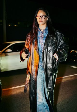 A photo of Rihanna wearing a denim jacket and jeans underneath a black leather trench coat.