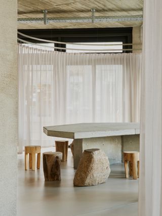 Interior view of JIGI Poke restaurant featuring light coloured floors, a concrete table, wooden seats, boulder seats and white curtains - designed by Vaust studio
