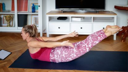 woman performing a dart pilates move on an exercise mat in a living room setting, wearing a pink vest and leggings.