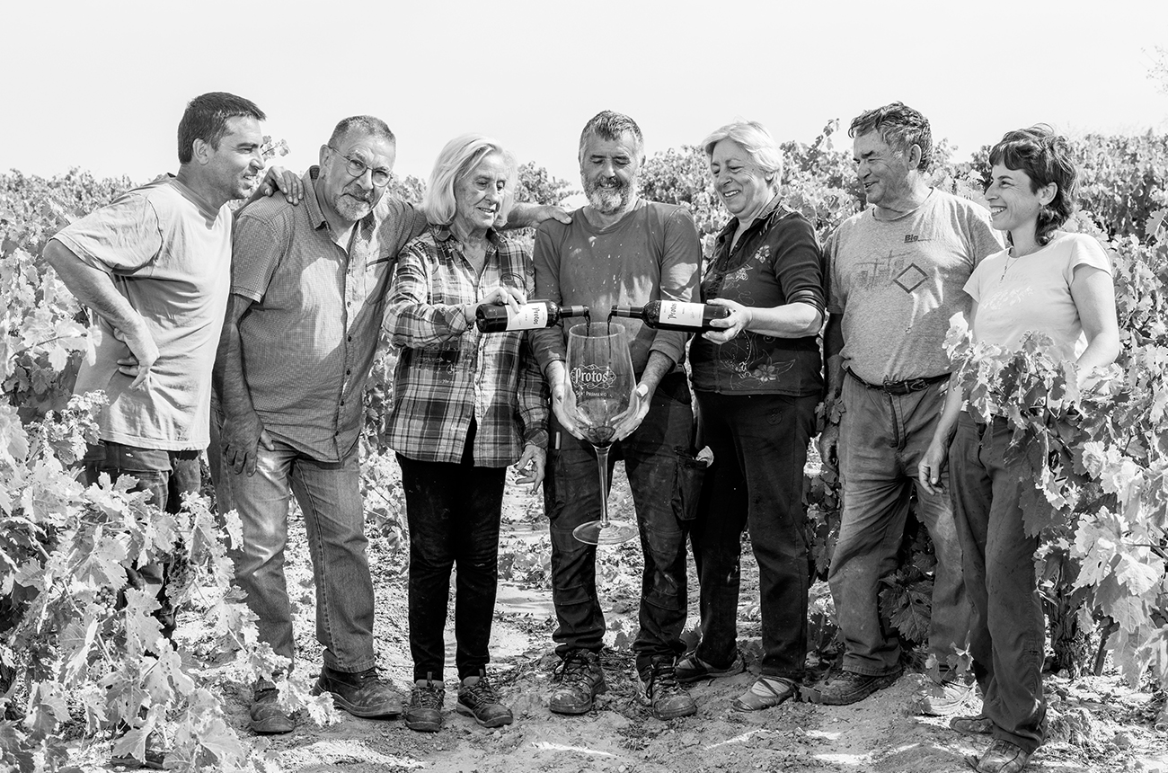Black and white photo of seven people standing between some vines, with the man in the middle holding a huge wine glass which says 'Protos' on it, and two people next to him pouring wine into it