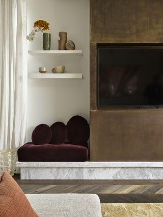 Image of a living room fireplace that is made out of a muted bronze-gold metal. The bottom ledge is a white marble and there is a little bench seat to the side of the fireplace that has oxblood red velvet upholstery.