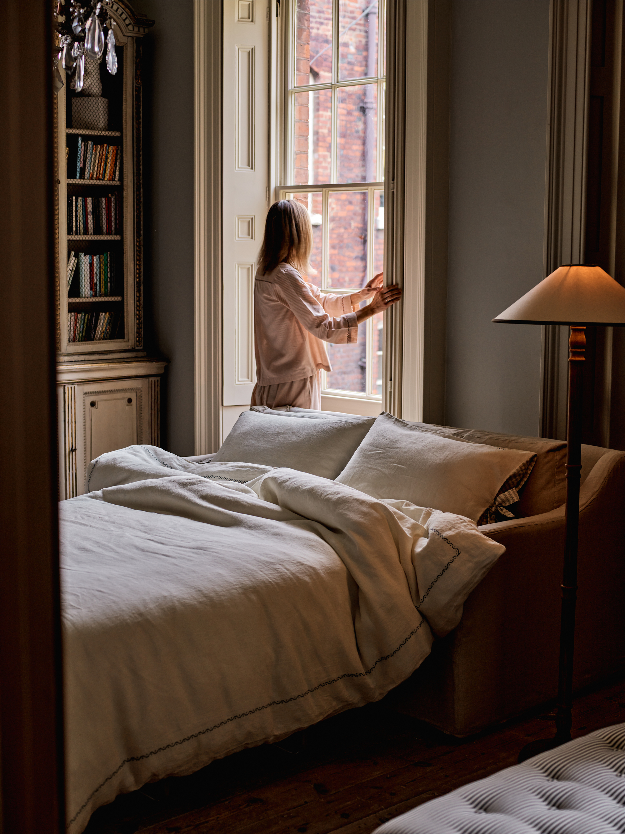 room with sofa bed folded out and styled like a bed, floor lamp, built-in bookshelf and woman standing in front of window, opening it