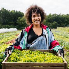 A smiling woman stands on a farm holding a crate of vegetables