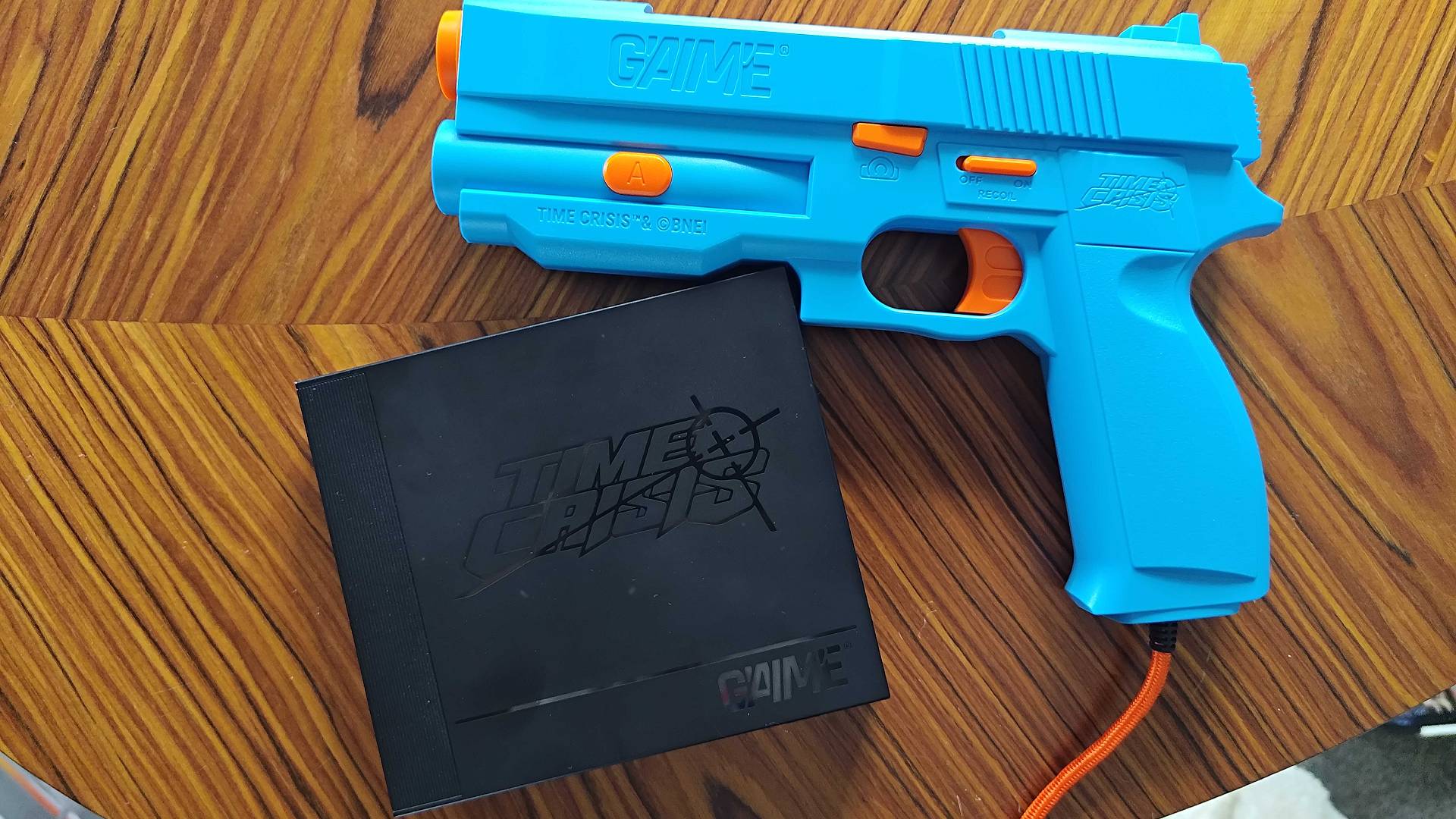 G&#039;AIM&#039;E Time Crisis console and blue lightgun on woodgrain desk.