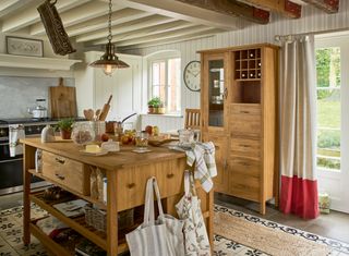 a wooden, country-style freestanding kitchen, with a tall boy, a wooden island full of ingredients and tote bags, and a lack age in the distance, on a tiled black floor with a huge rug
