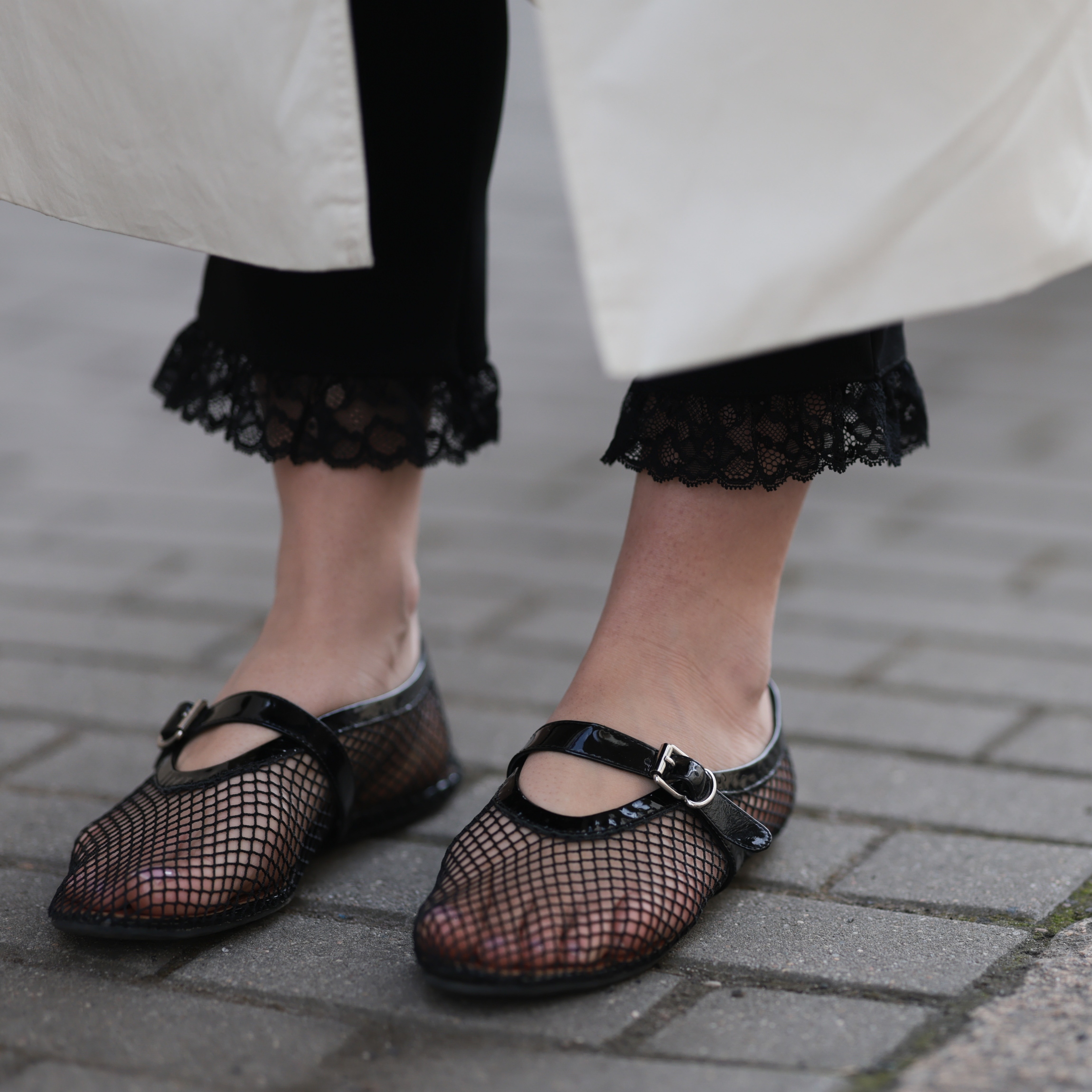 Close-up of black mesh Mary Jane flats styled with lace-trimmed black pants and a white coat.