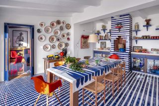 The exteriors of an Italian villa painted in color block shades of red, blue, and white stand out against the dense vegetation visible in the background and the terracotta tiled flooring of the swimming pool in front of it.