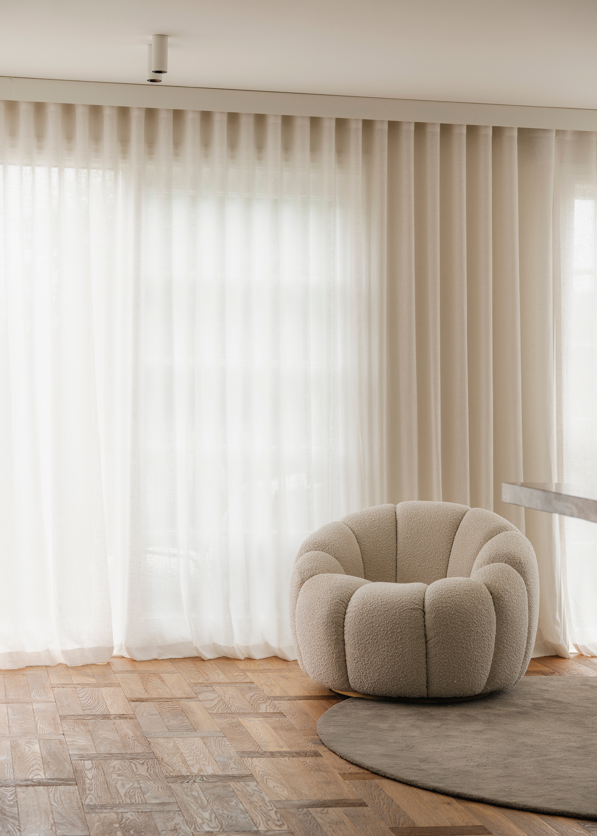 A pared-back minimalist living room with wood flooring, a textured sofa seater, sheer curtains and a circular rug
