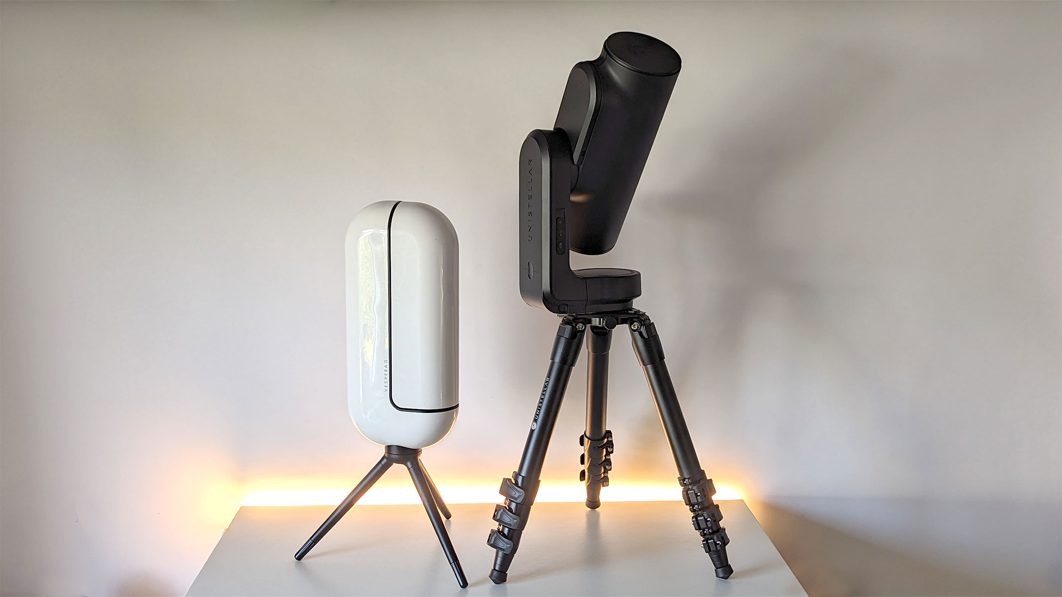 The Vaonis Vespera 2 and the Unistellar Odyssey Pro side-by-side on a white table and background, demonstrating their size difference.