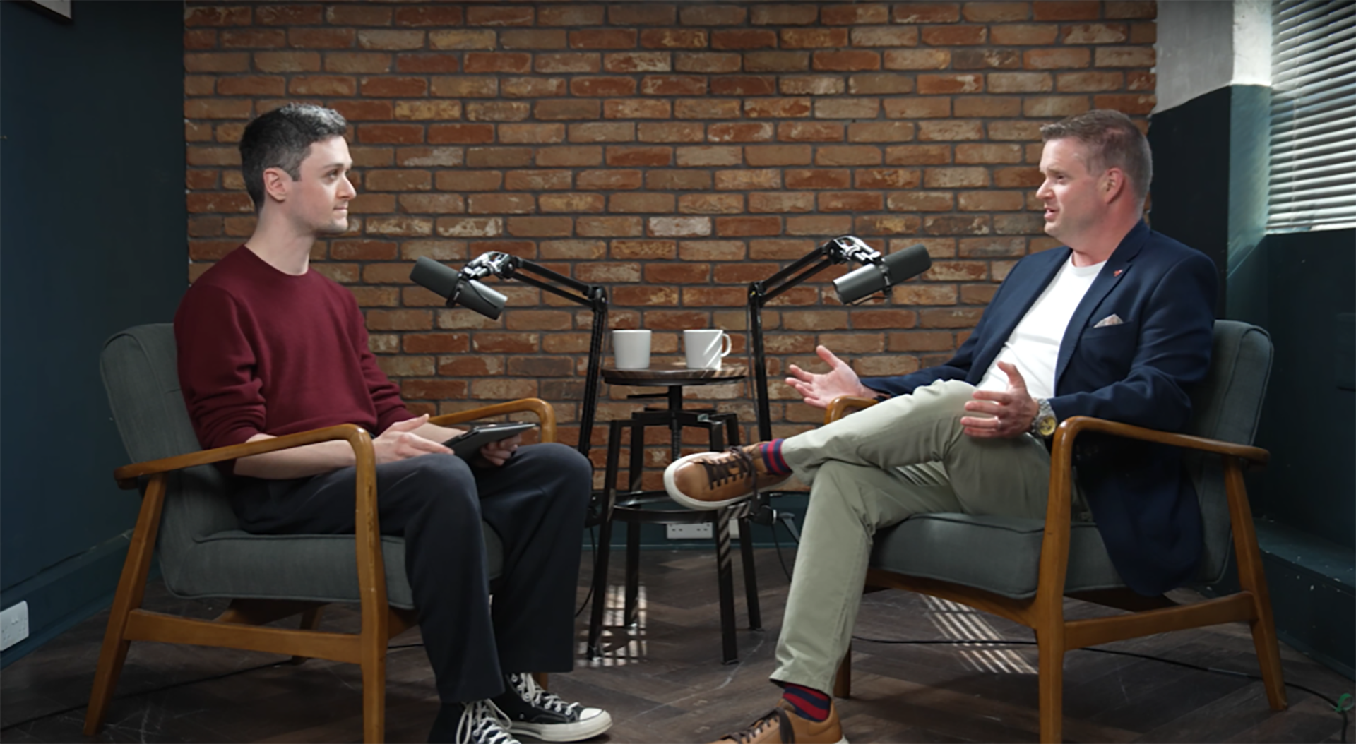 ITPro Features Editor Rory Bathgate and Red Hat Director of Business Value Practice Ed Hoppitt facing each other in a room with one wall of exposed brickwork and the other two visible walls painted dark teal. Rory is listening while Ed is talking
