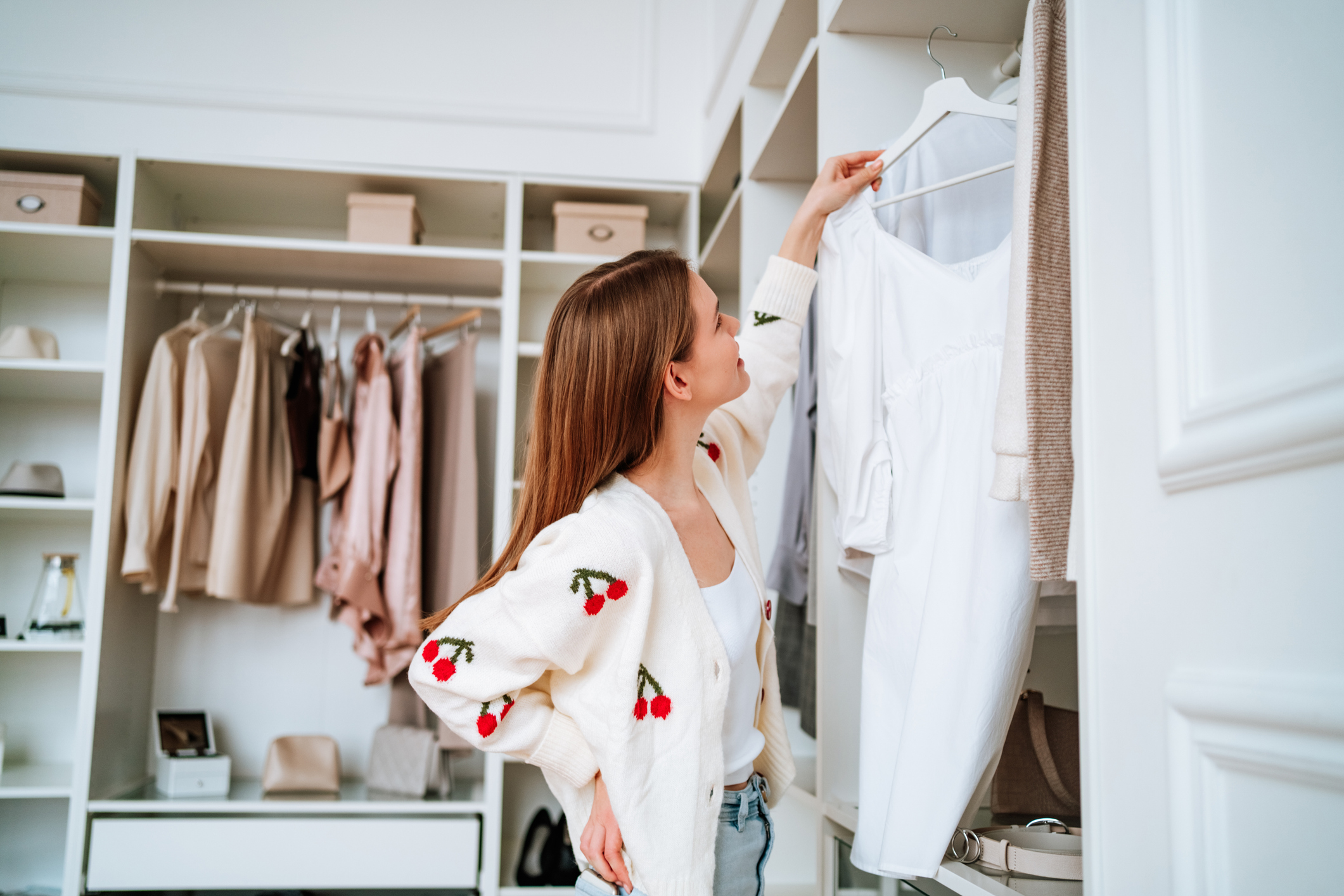 A woman wearing a white cardigan with cherries on, looking at clothes in her walk-in wardrobe.