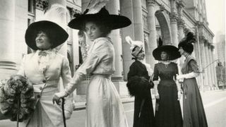Women in Edwardian-style dresses and large feathered hats pose with parasols on a city street in front of a neoclassical building. Is this helpful so far? Ask ChatGPT