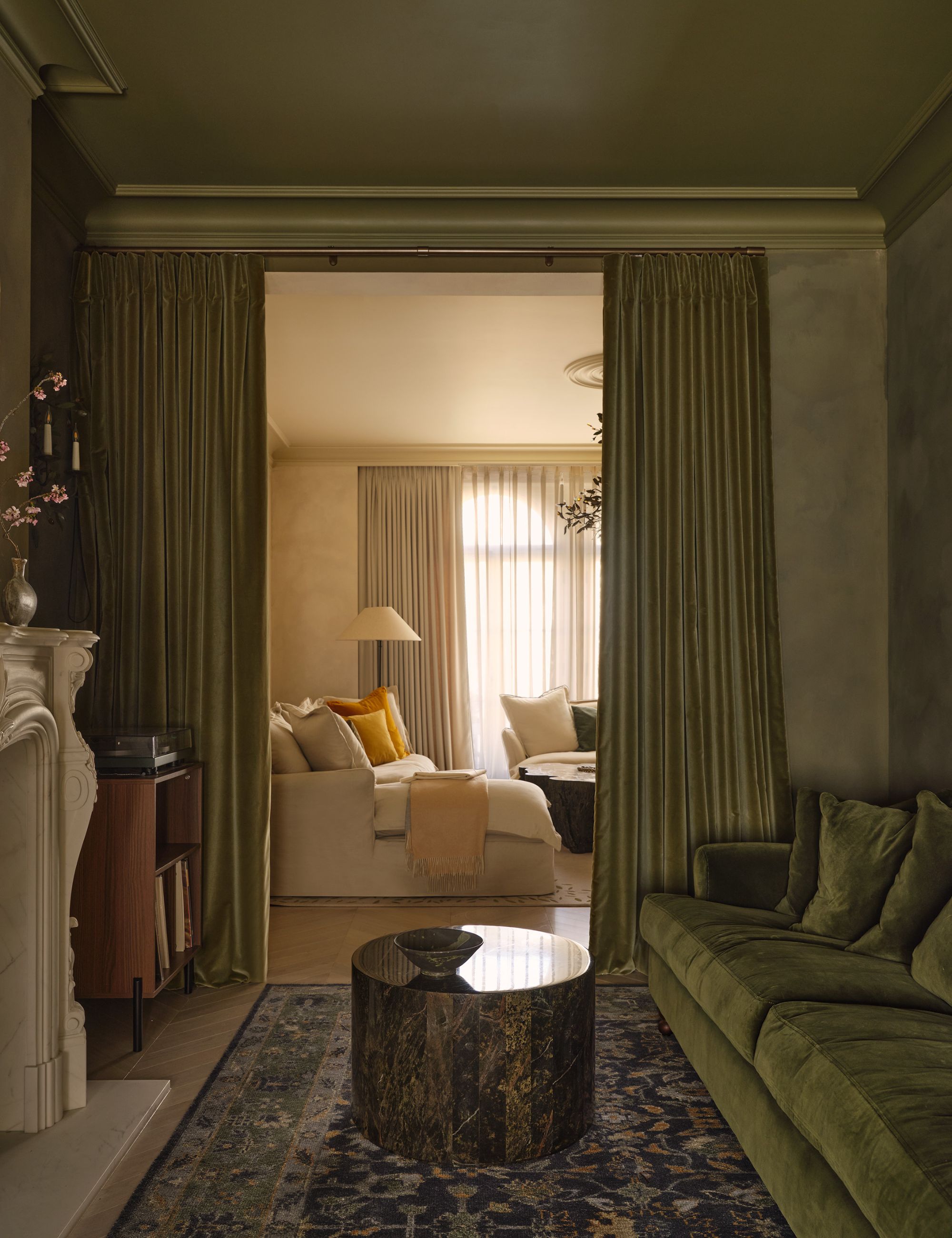 Tonal green front room with lime washed walls, circular table, pattered rug, marble fireplace and curtains