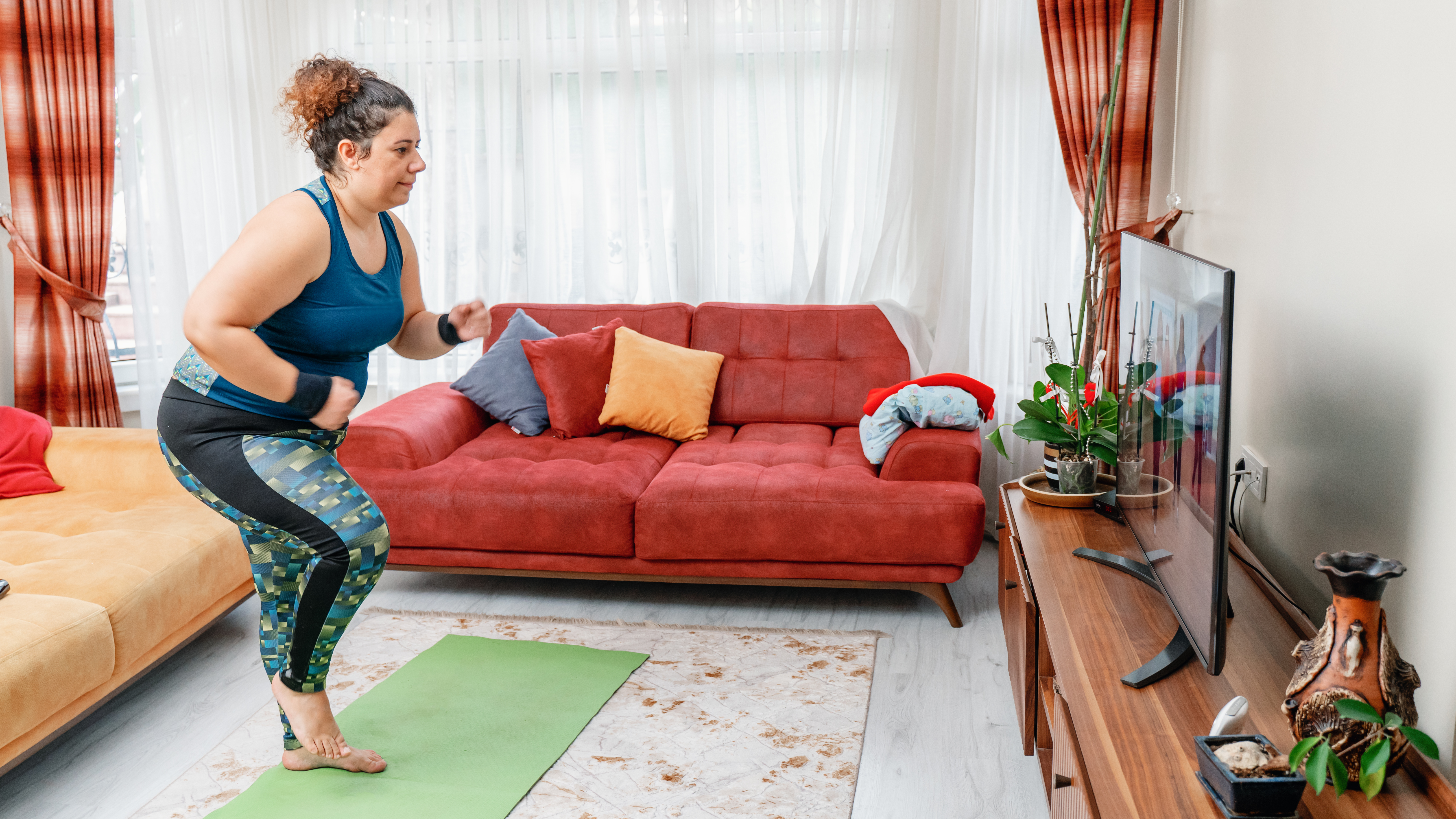 A woman exercises at home on a mat while watching a television. She is marching on the spot. 