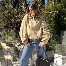 Sofia Richie Grainge poses on a balcony wearing a light beige suede jacket, blue vintage Levi jeans and satin pointed-toe heels. She holds a black leather bag in her hand and wears black sunglasses.