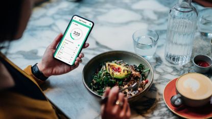 Woman holding a phone with calorie counter app, next to bowl of healthy avocado salad, representing how to lose a stone in a month