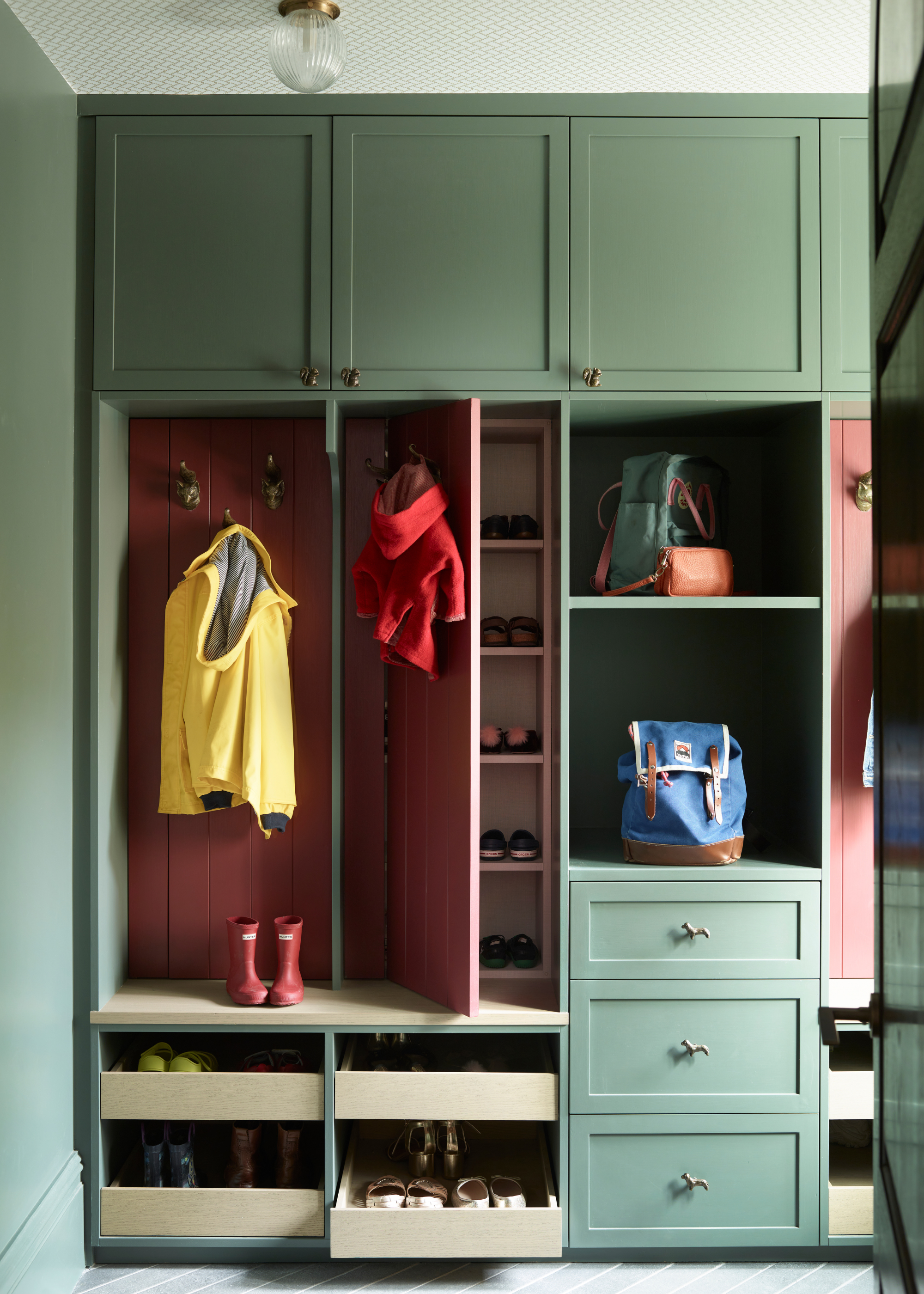 A cleverly designed dark pin and green storage unit in a mudroom which features hidden shoe storage behind one of the panels
