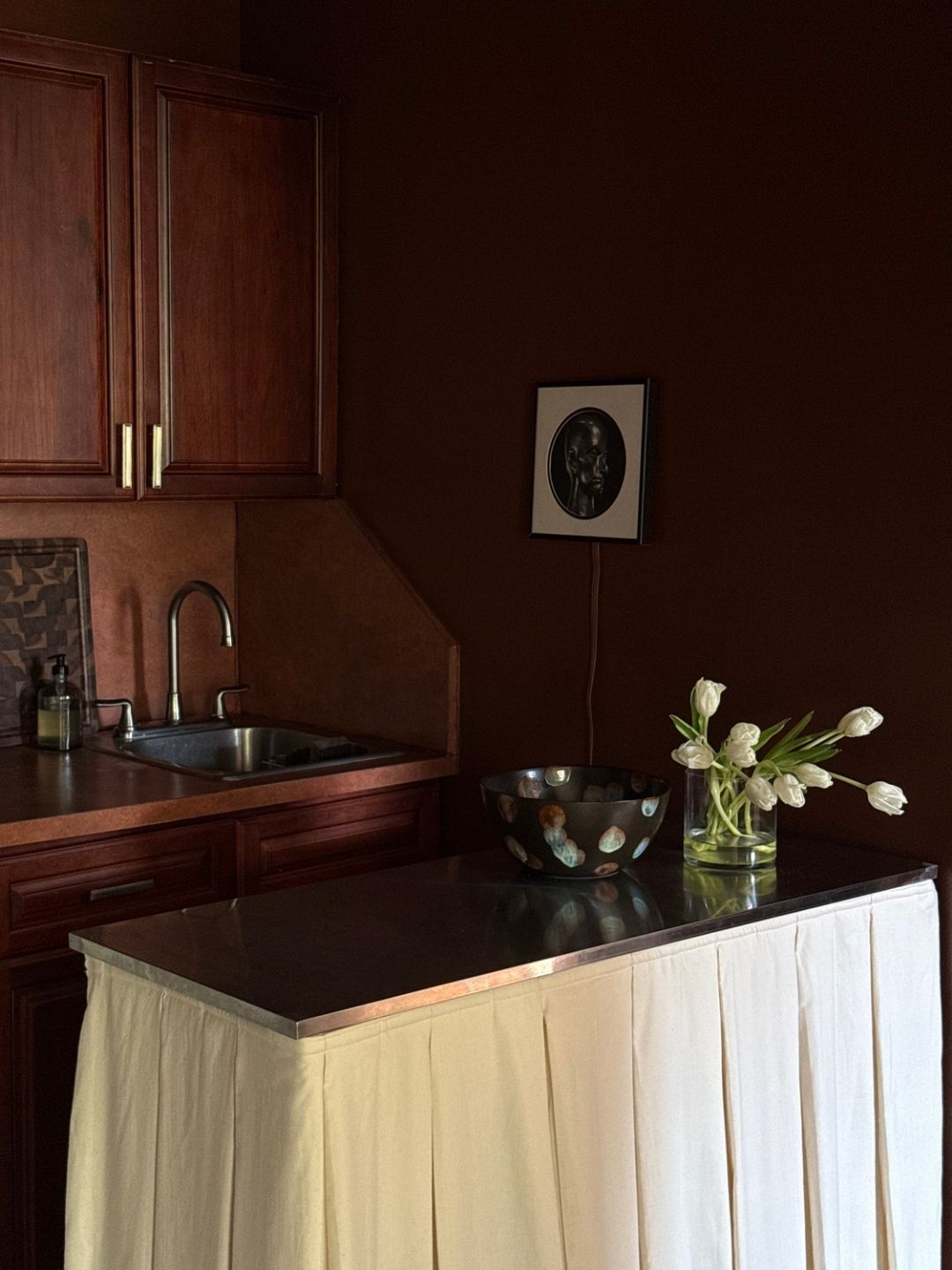 Image of a dark reddish-brown kitchen with a small kitchen island that has a chrome top and a white box-pleat skirt.