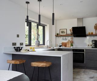 Contemporary kitchen with a minimalist aesthetic, featuring black pendant lights, a grey countertop, and wooden bar stools.