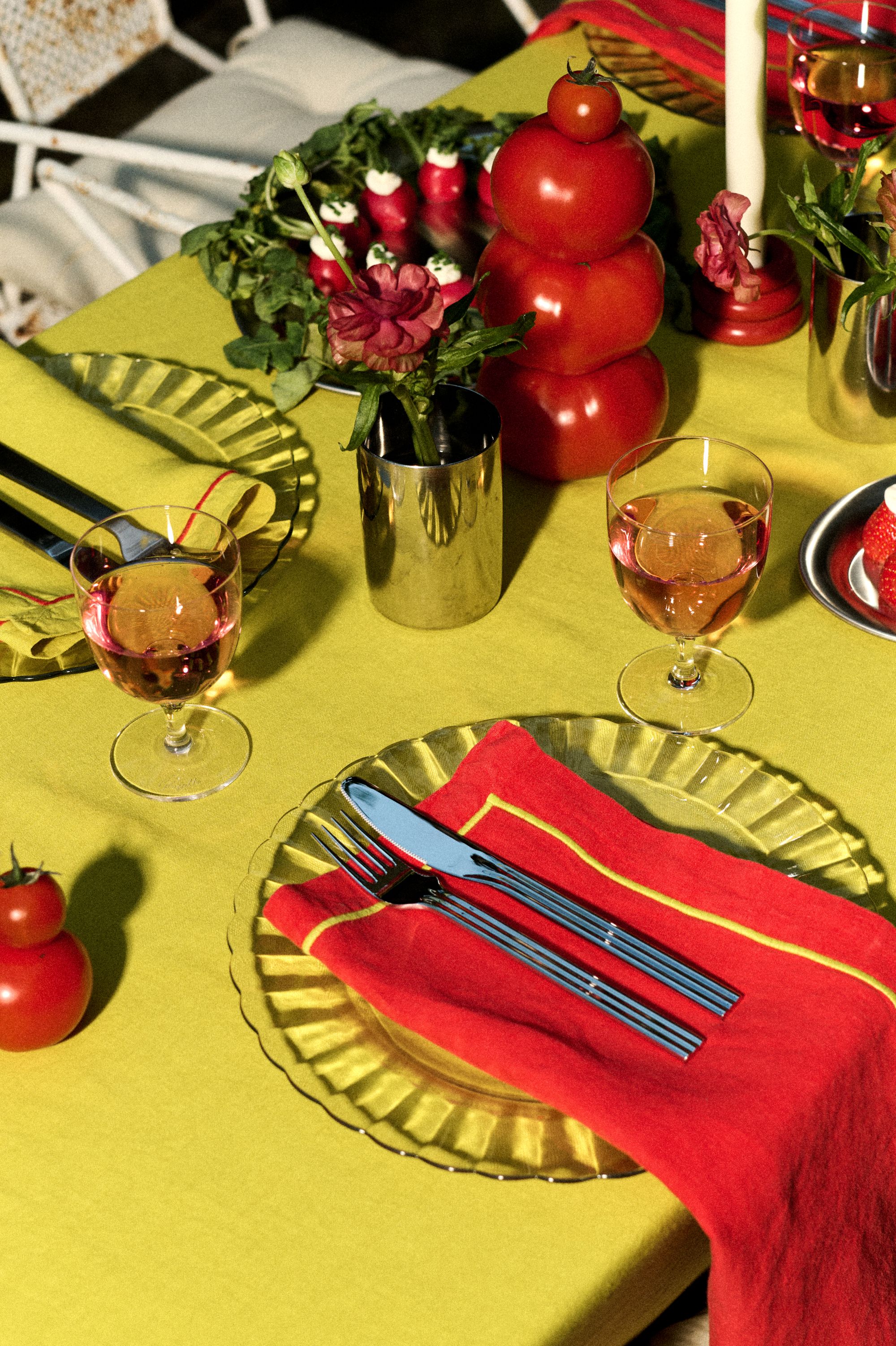 a table with a green table cloth, clear plates, silver serveware, a red napkin, wine glasses, and tomatoes for decoration