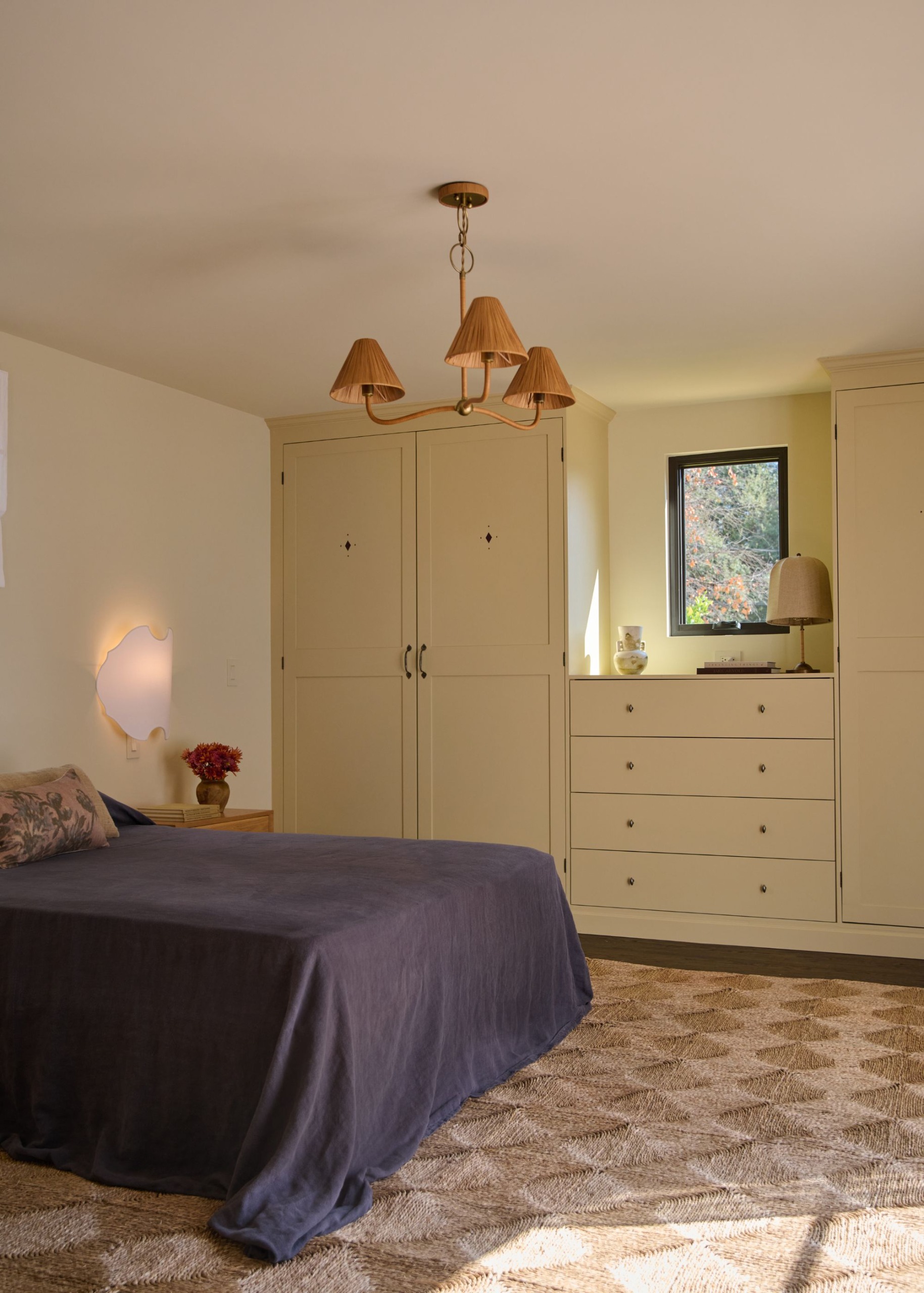 A modern bedroom with off-white walls, a wall of butter-yellow joinery with a small window in the middle. A jute-patterned rug, a low bed, and a rattan ceiling light fixture.