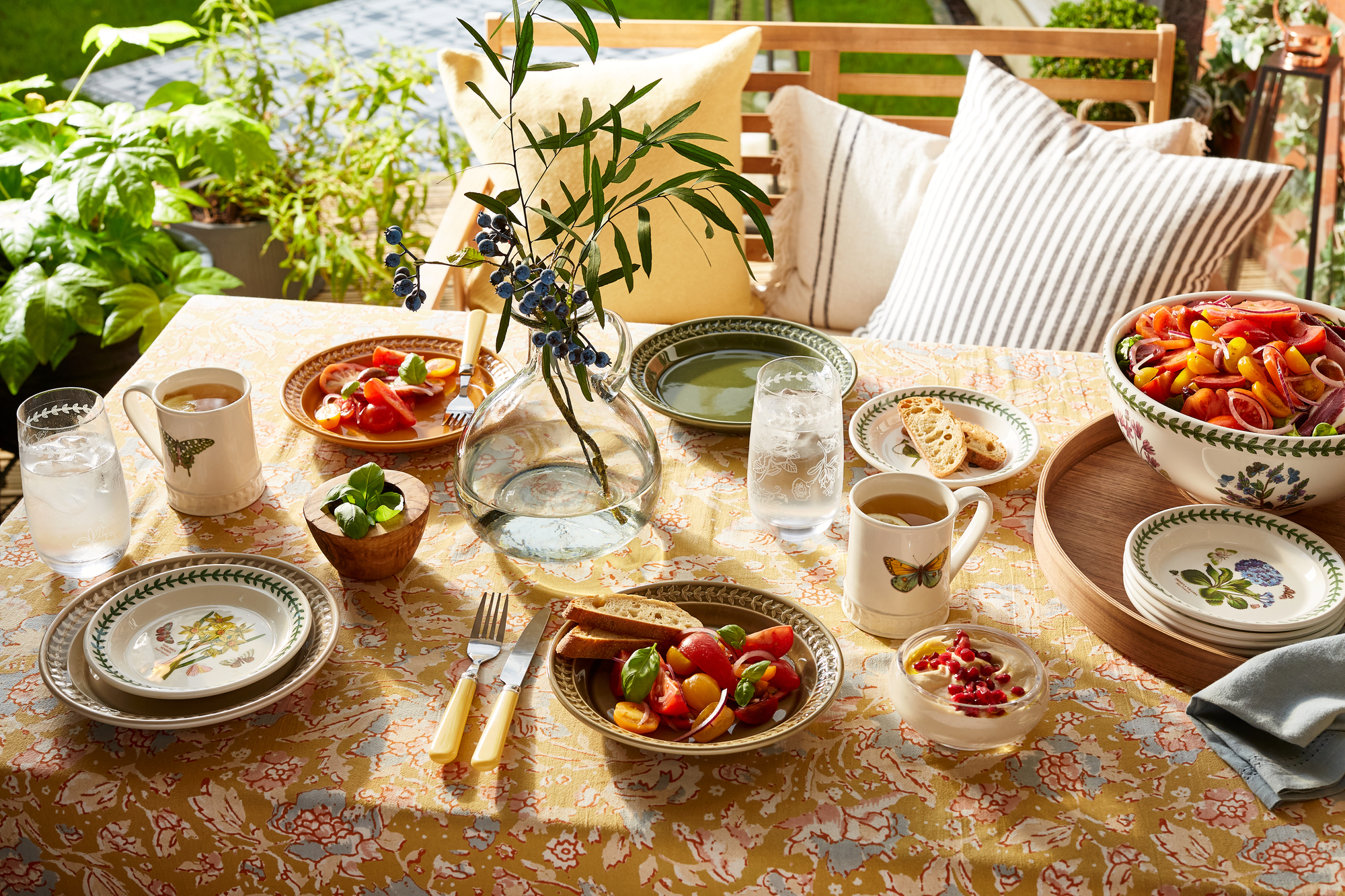 An outdoor dining table laid with a patterned tablecloth, bowls and plates, and a vase of foliage