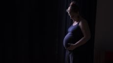 A pregnant woman in a purple tank top looks down at her belly standing in front of a black background