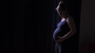 A pregnant woman in a purple tank top looks down at her belly standing in front of a black background