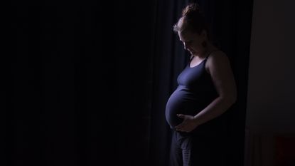 A pregnant woman in a purple tank top looks down at her belly standing in front of a black background