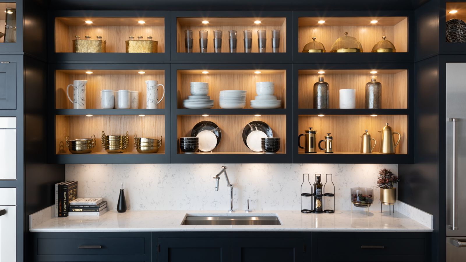 A sleek kitchen with dark cabinetry, open shelving and warm accent lighting, highlighting glassware and ceramics above a pale stone worktop.