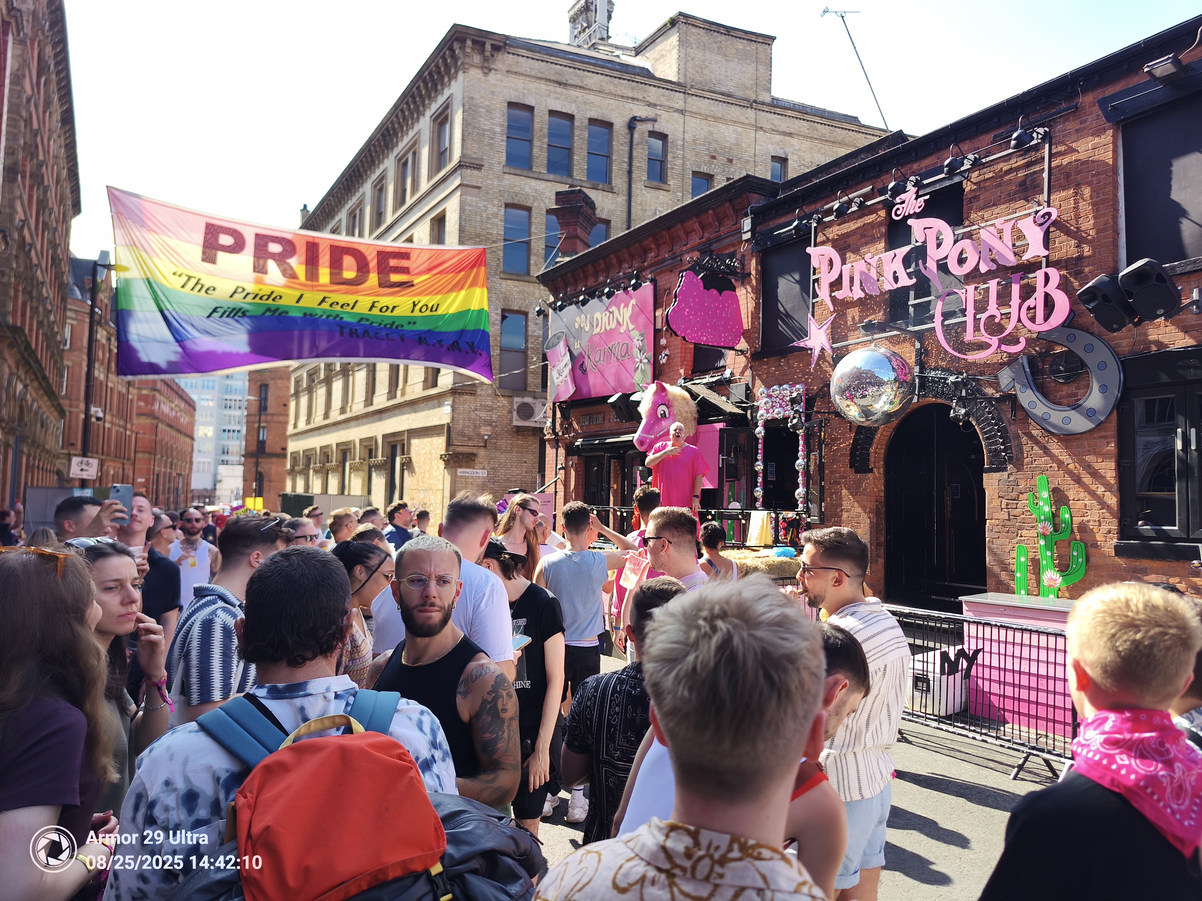 A diverse crowd gathers on a city street adorned with a large rainbow flag that reads "PRIDE," with a building in the background featuring a pink sign that says "The Pink Pony Club."