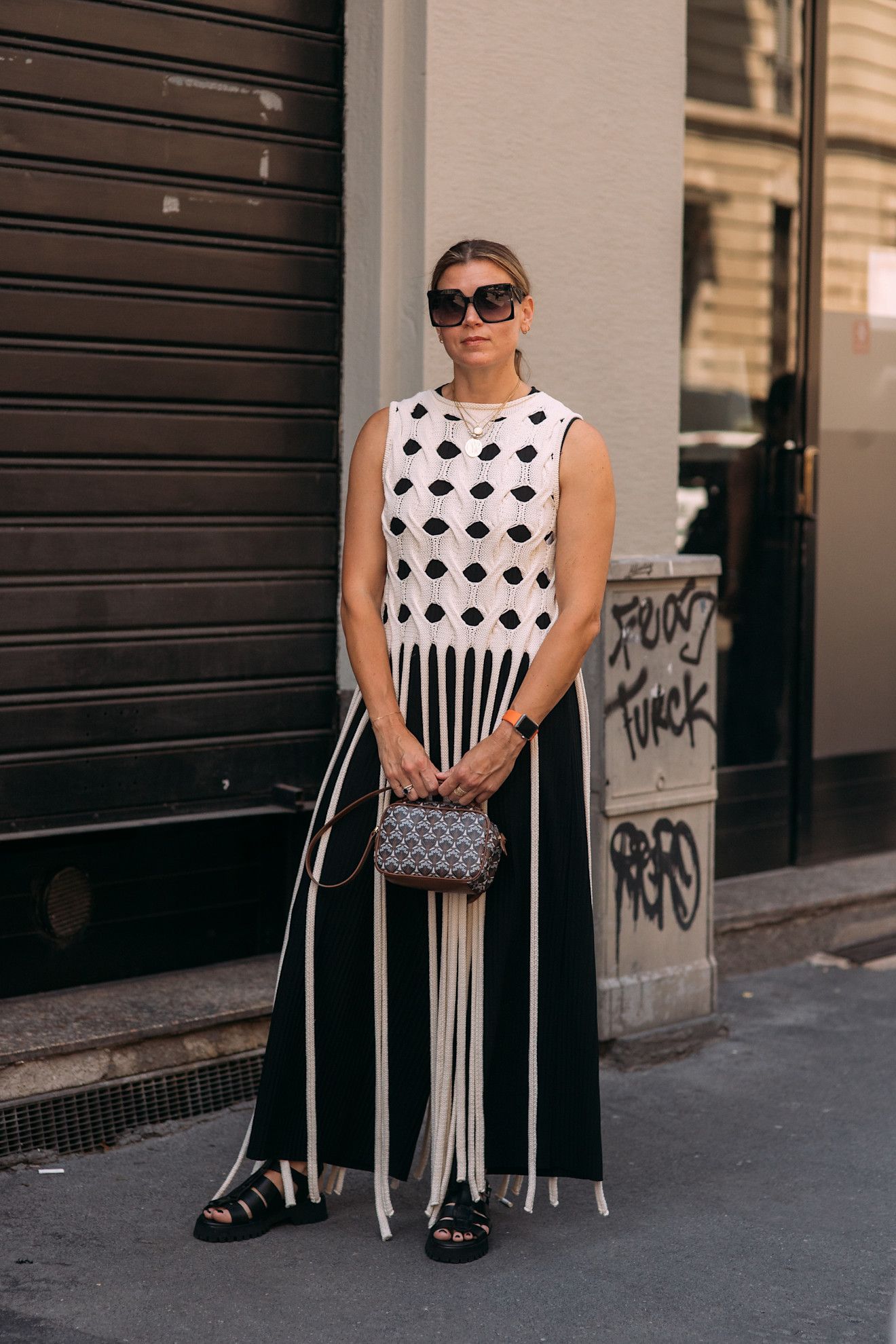 Menswear SS26 showgoer wears an open-weave sleeveless top with floor-length tassels layered over a black skirt with black fisherman sandals.