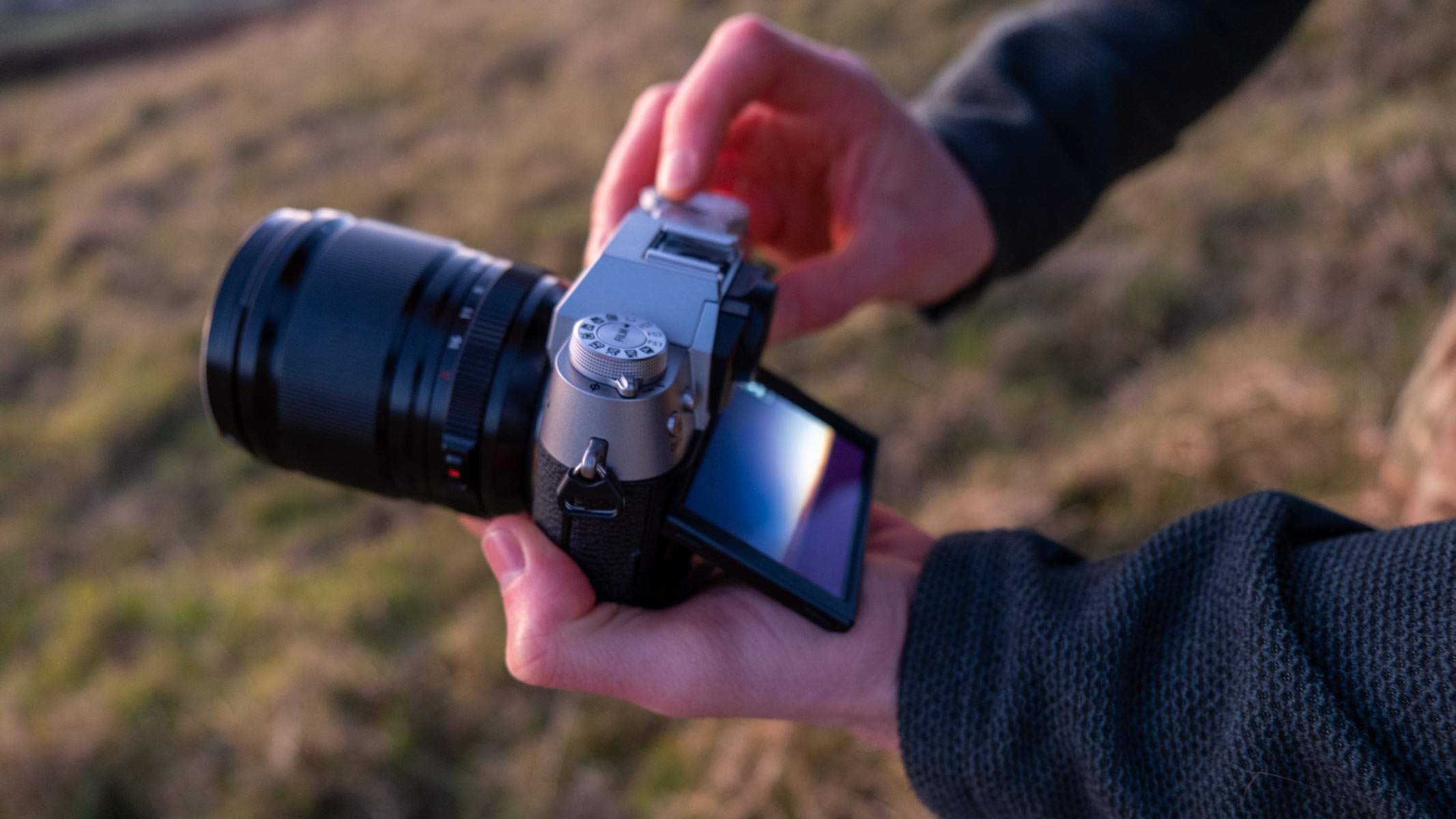 Hands holding the Fujifilm X-T50, with its tilted LCD screen showing a sunset landscape