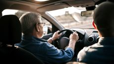 An elderly man drives a car with a younger man in the passenger seat.