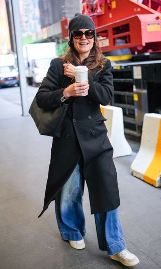 A photo of Katie Holmes bundling up in a black longline coat, jeans, Autry sneakers, and a black tote bag.