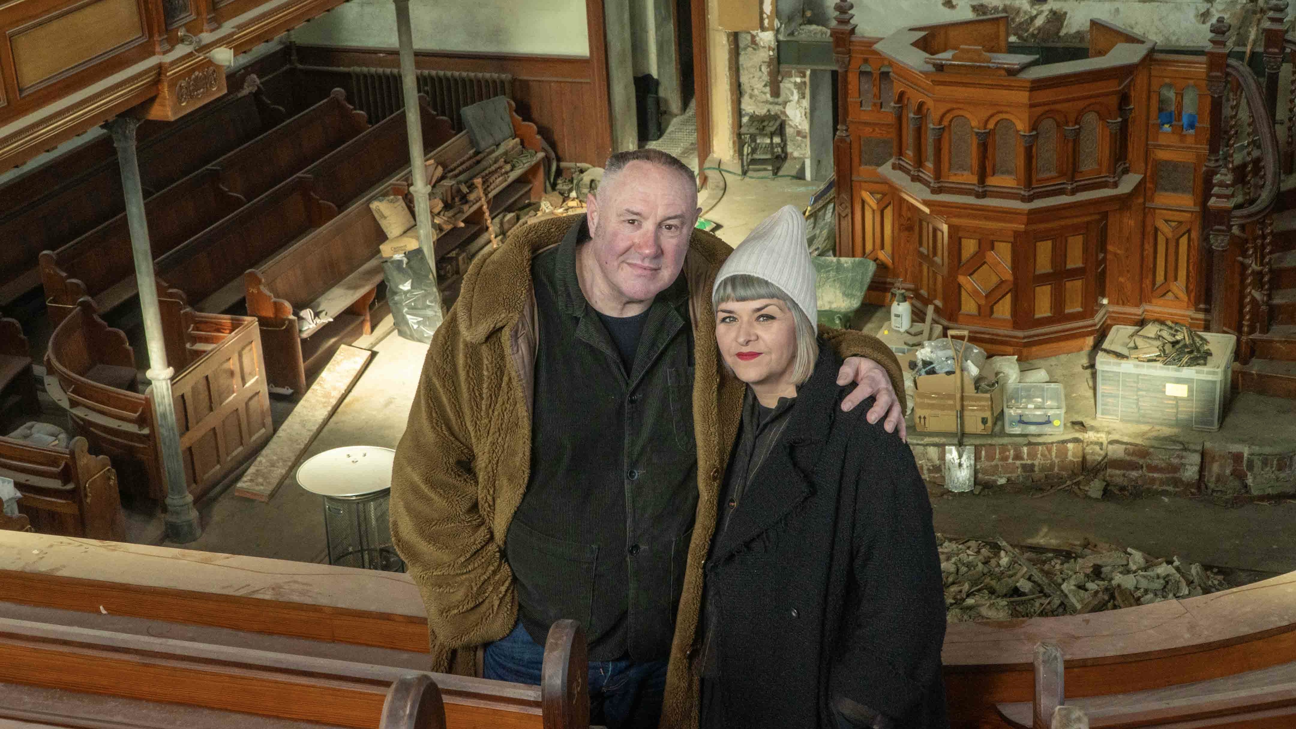 Keith Brymer Jones and Marj Hogarth in warm coats stand against a backdrop of their chapel complete with pulpit in Our Welsh Chapel Dream