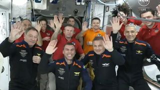 11 people in flight suits wave to the camera aboard the white-walled international space station.