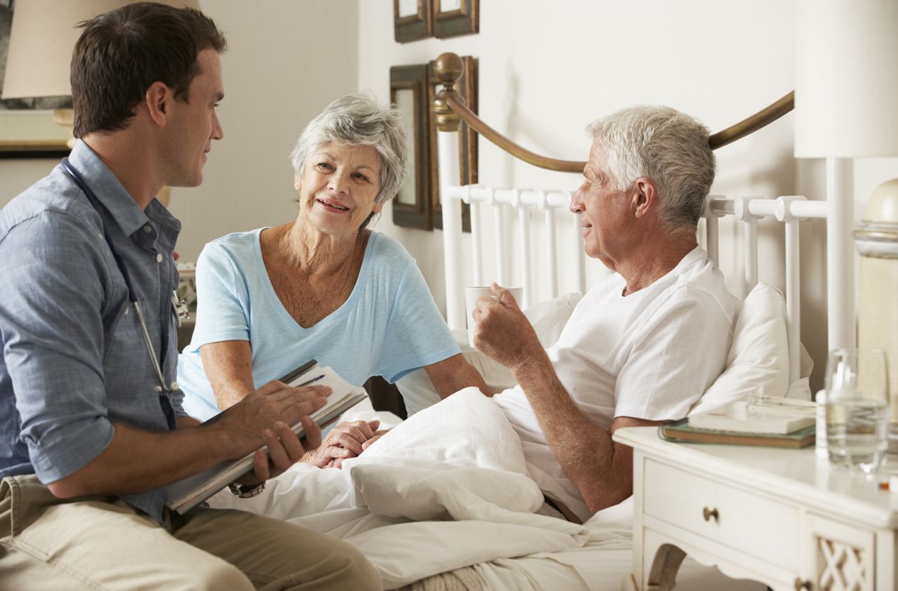 Doctor On Home Visit Discussing Health Of Senior Male Patient With Wife In Bedroom.