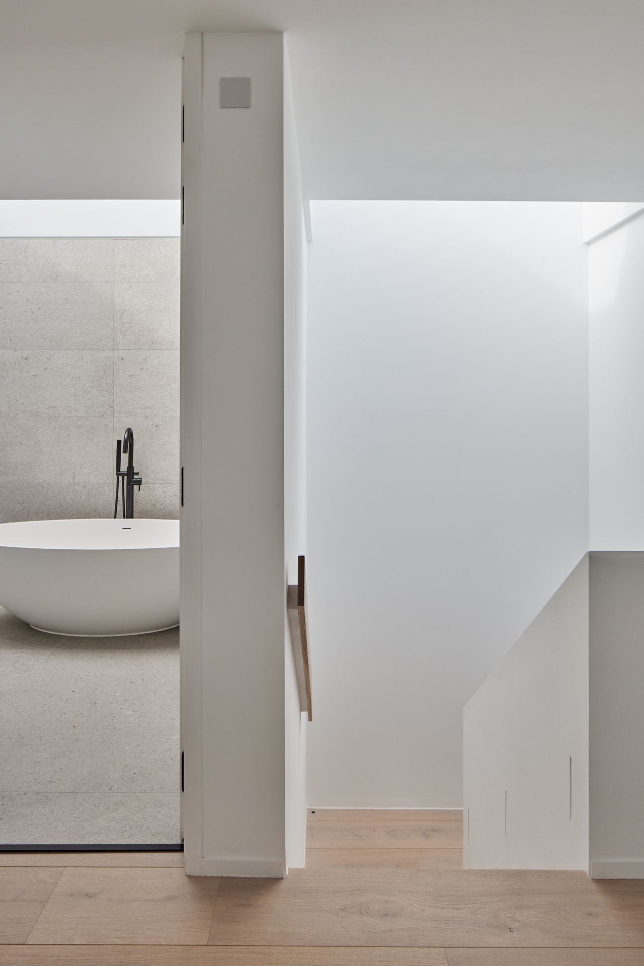 A view of a modern landing with one doorway open to reveal a minimalistic bathroom and a stairway leading downstairs