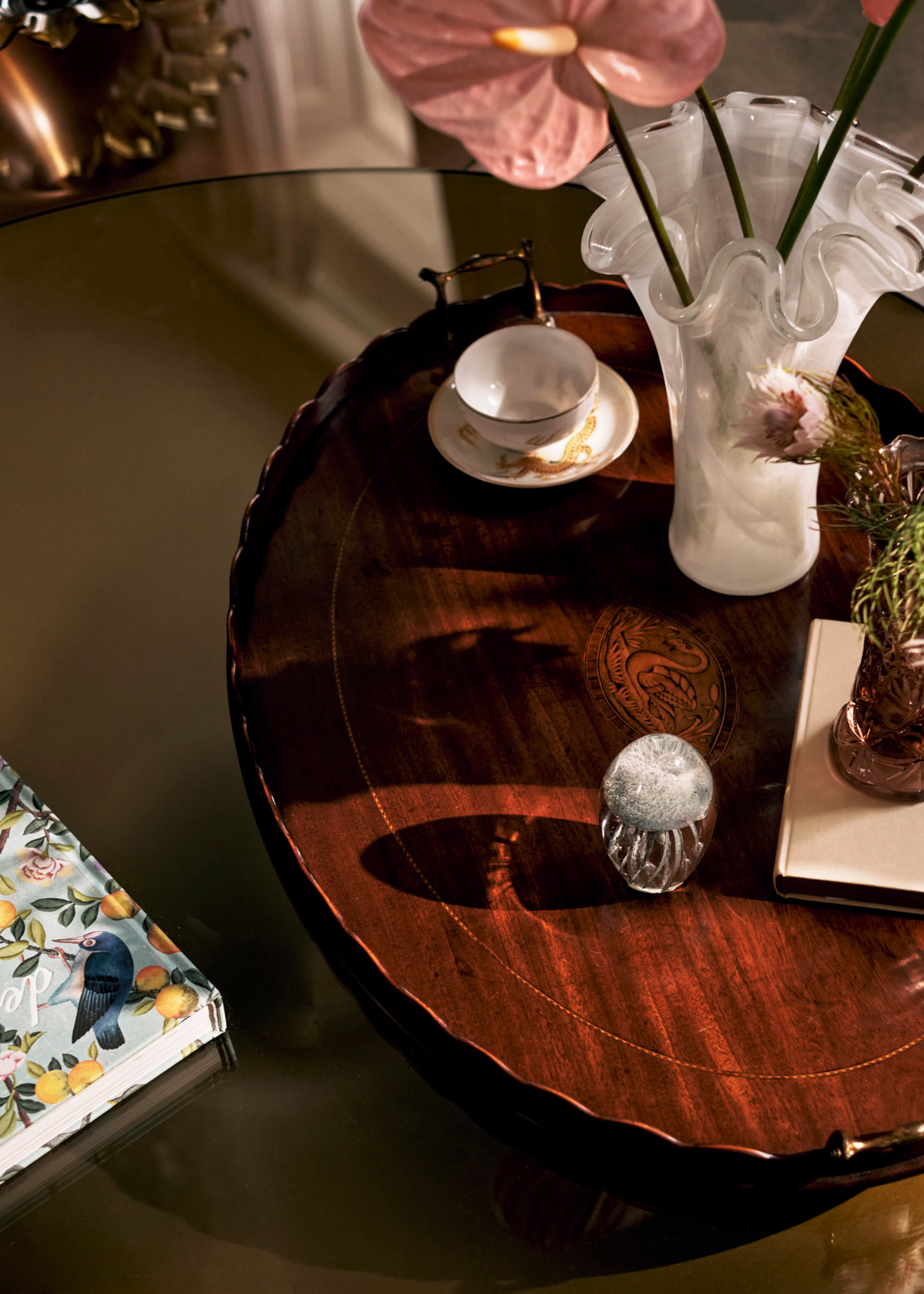 A close-up of a glass coffee table with a tray holding a vase of pink anthuriums, a bowl, and a planter