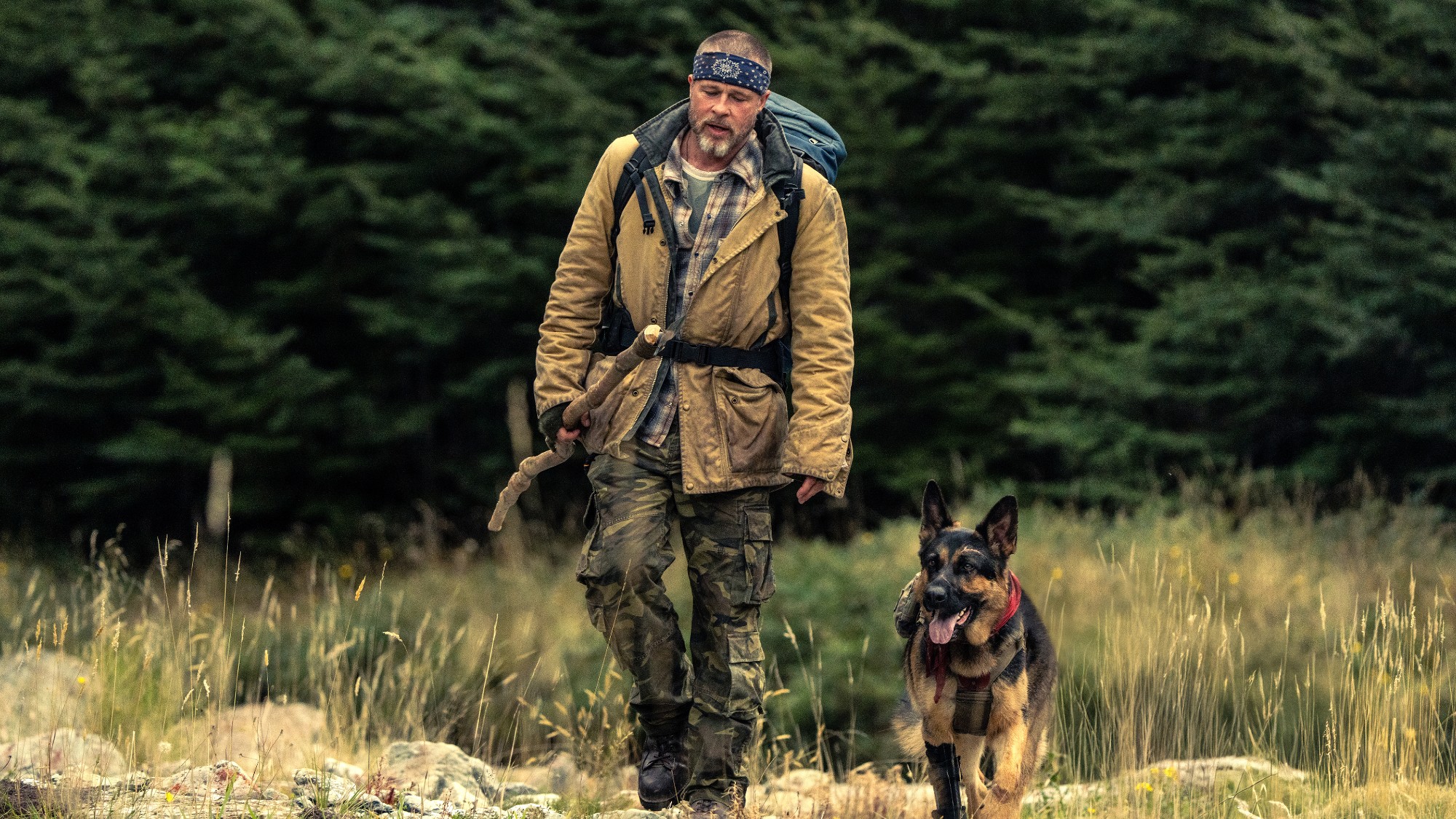 Brad Pitt in the wilderness with a dog. He is carrying a stick and wearing a tan jacket and a blue bandana around his head in Heart of the Beast.