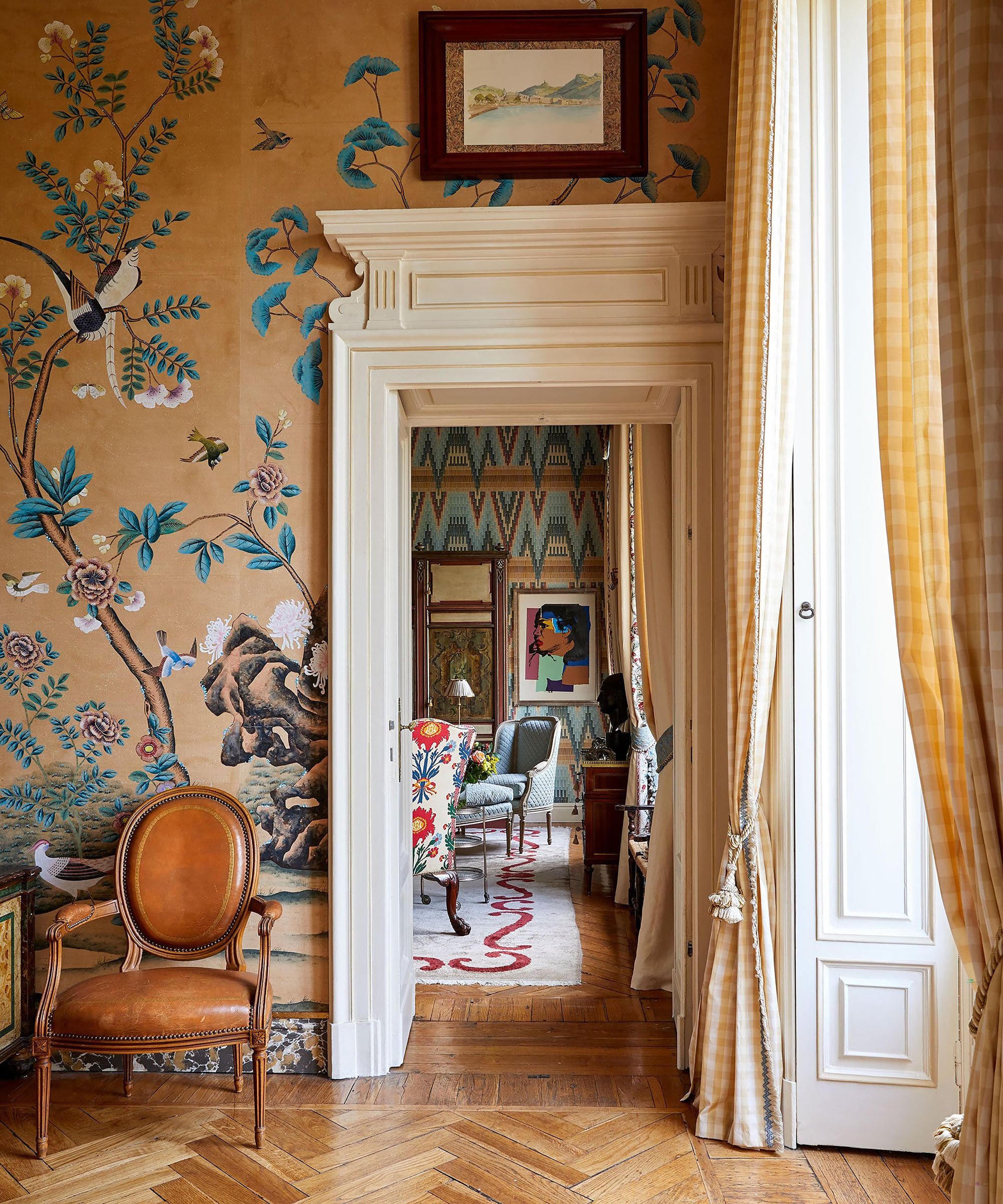 Tall living room with ornate door frame and striking orange wallpaper. The floor is original parquet, there are long floor to ceiling windows dressed with checkered curtains