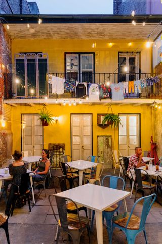 A yellow-tinted historic building houses a laid-back eatery and drink hotspot with white square tables, iron blue or unfinished chairs, and fairy lights bringing the space to life.