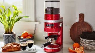 A red KitchenAid Burr Coffee Grinder girnding coffee next to two shots of espresso and a peeled orange