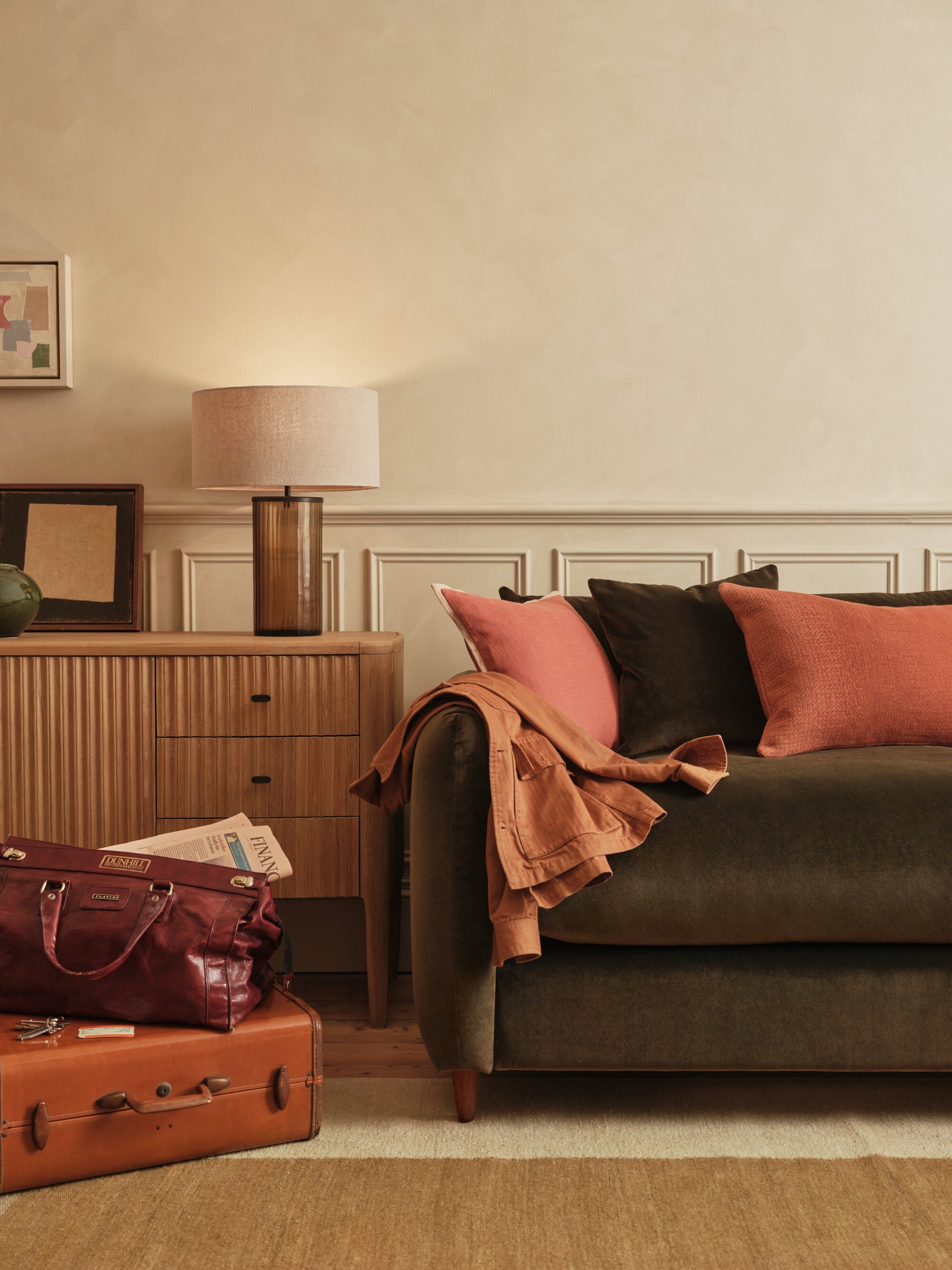 Image of a dark green, velvet sofa bed in a beige living room. There are pink and green pillows on the sofa and a fluted side table with an amber glass lamp next to the sofa. There are two suitcases stacked in the corner.