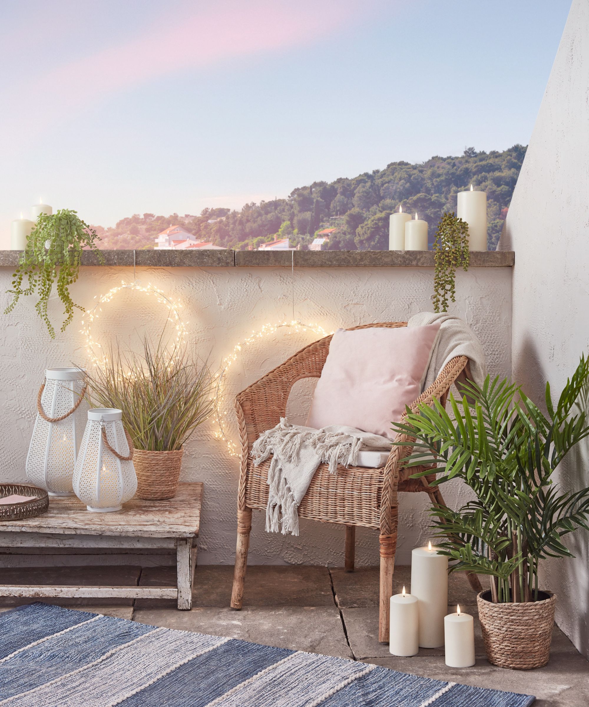 Cosy balcony setup with rattan chair, soft cushions, candles, lanterns and string lights, styled with potted plants and a view of tree-covered hills.