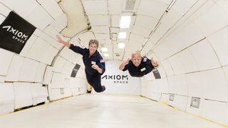 view of an airplane interior with two astronauts floating. the words 'axiom space' are on the wall and at back
