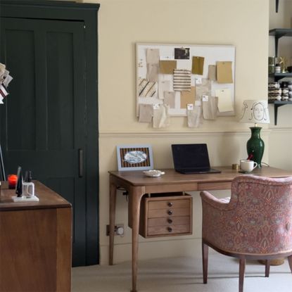 Neutral home office with wooden desk, patterned upholstered armchair and pinboard on wall above desk showing fabric samples