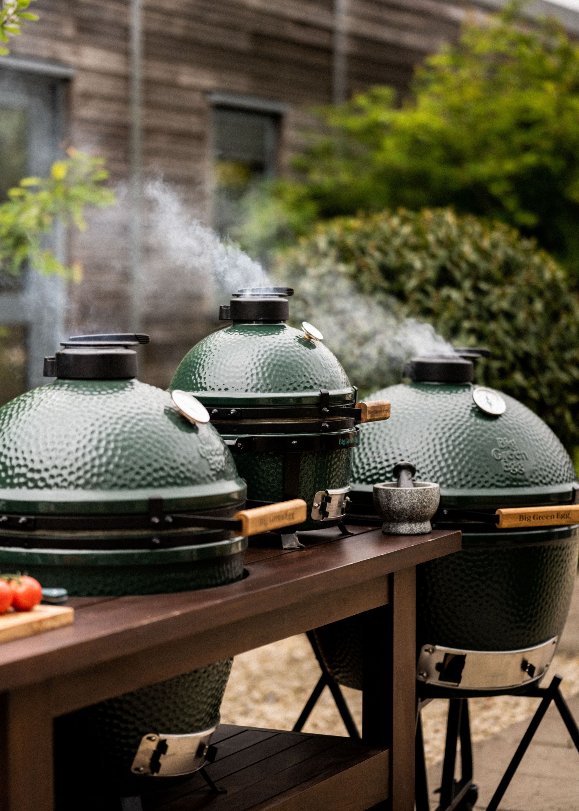 Three large Big Green Egg grills in a garden surrounded by plants