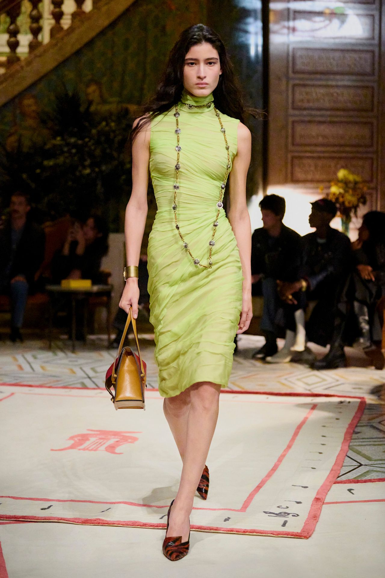 A Versace model wearing a layered silk slime green high-neck midi dress styled with a long silver and gold chain necklace, a brown structured handbag, and zebra print pointed pumps.